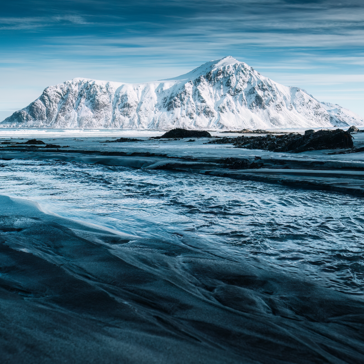 Snowy mountain peak with a rocky shoreline and blue water.